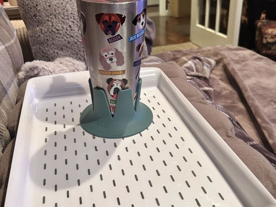 A sage-green drink base holding a stainless tumbler wrapped in a dog-print design, standing on a white slotted serving tray on a soft grey blanketed surface in a living room