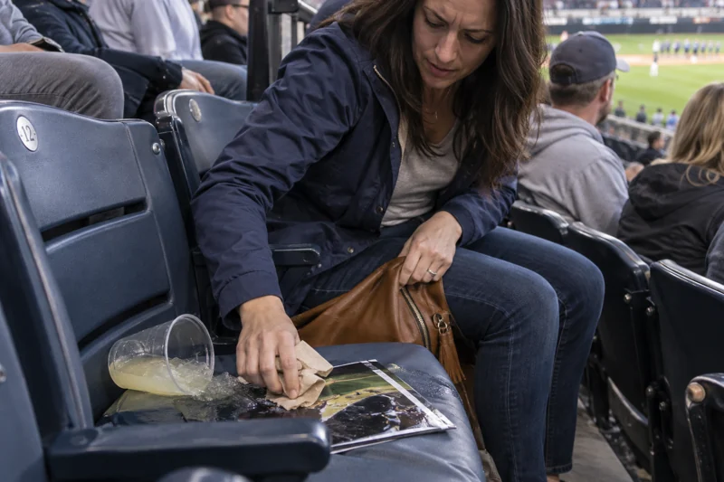 Reaching down for bag tips drink on seat beside them; liquid wets stadium cushion and program. AI illustration.